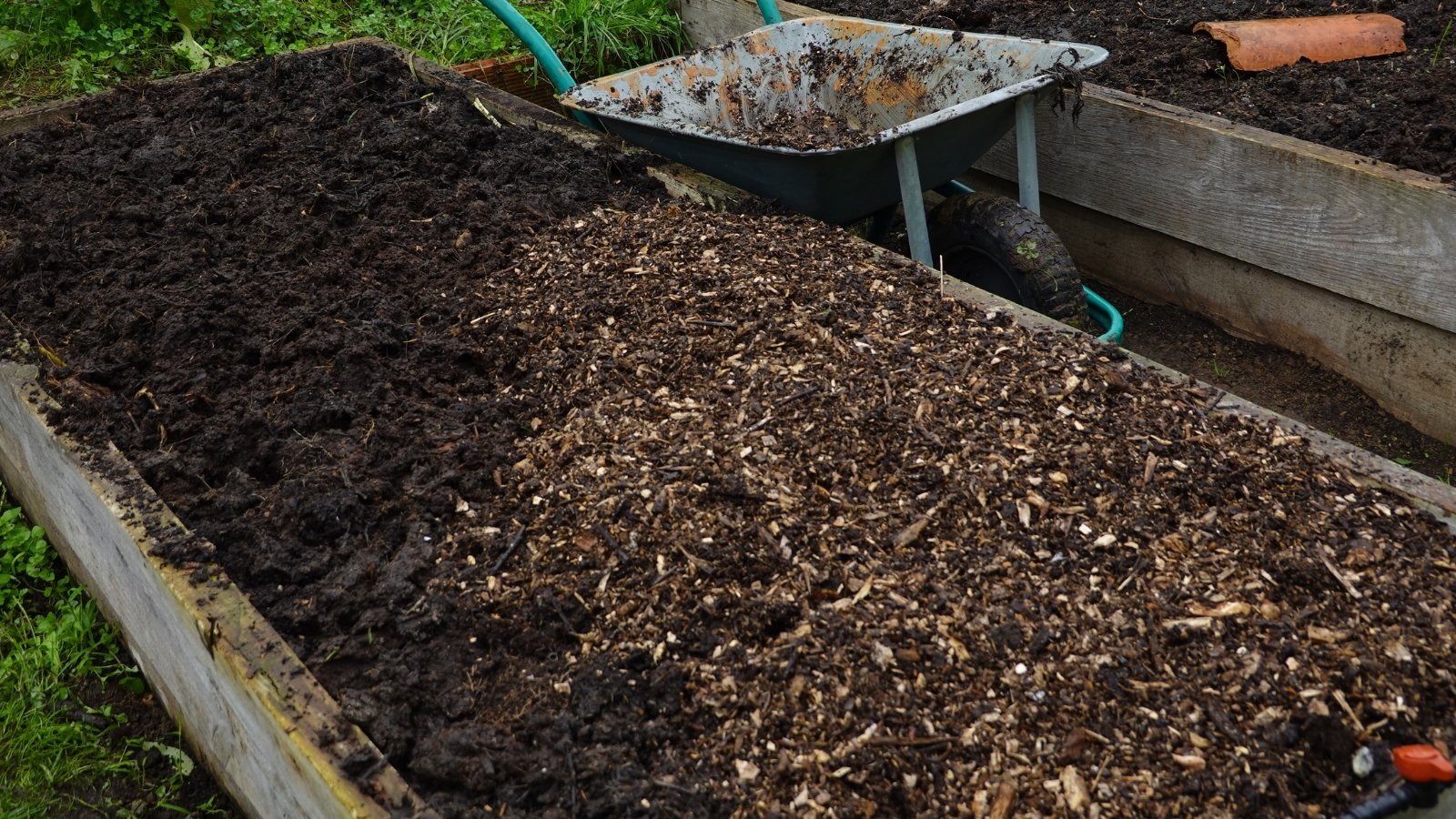 A raised rectangular soil bed covered with a thick layer of dark mulch, freshly spread and tilled, sits near a wheelbarrow on a bare dirt ground with patches of green foliage nearby.