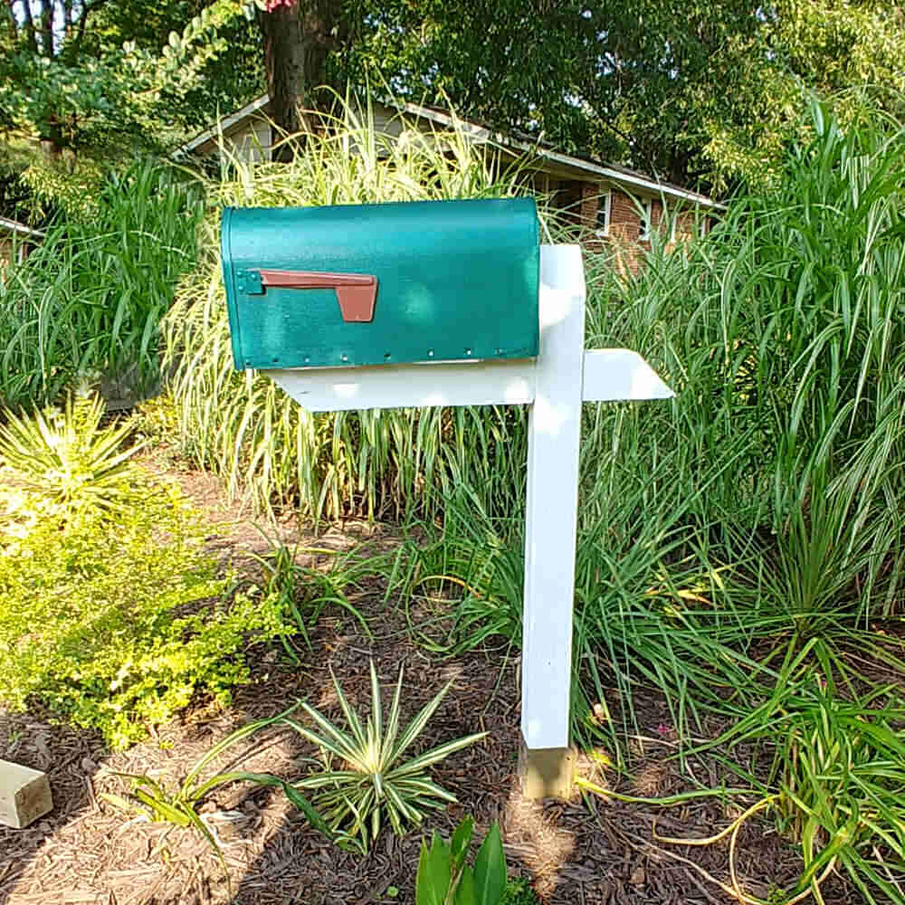 Green mailbox on a white post in a garden setting.